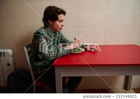 Teenage Boy Eating Salad at Red Table in Simple Kitchen, Quiet Home Moment Teenage Boy Eating Salad at Red Table in Simple Kitchen, Quiet Home Moment 133149823