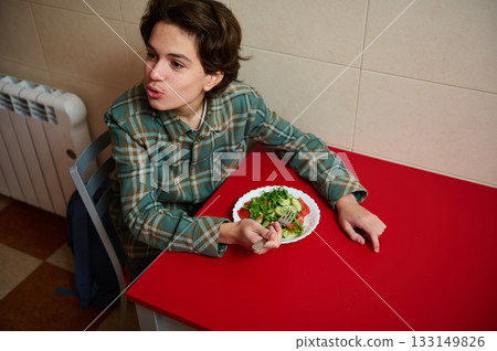 Teenage Boy Eating Salad At Red Table In Kitchen, A Glimpse Of Gen Z Daily Life Teenage Boy Eating Salad At Red Table In Kitchen, A Glimpse Of Gen Z Daily Life 133149826