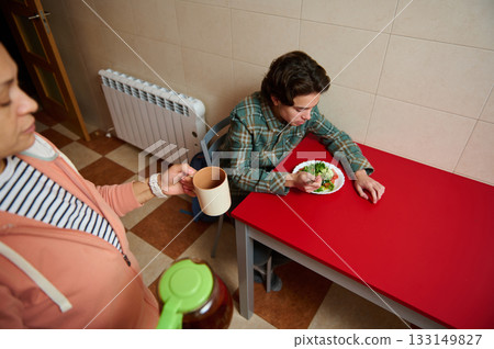 Teenage Boy Eating Salad at Red Table While Mom Serves Coffee in Cozy Kitchen Teenage Boy Eating Salad at Red Table While Mom Serves Coffee in Cozy Kitchen 133149827