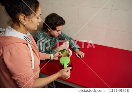 Mother Pouring Drink While Teen Boy Eats Salad At Red Table In Kitchen Mother Pouring Drink While Teen Boy Eats Salad At Red Table In Kitchen 133149828