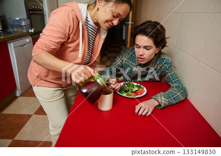 Mother Pours Juice For Teen Boy At Red Table Lunch In Kitchen Scene 133149830