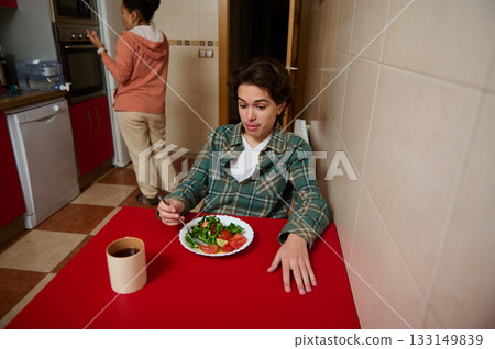 Teenager Enjoying Salad at Red Kitchen Table With Friend in Background Teenager Enjoying Salad at Red Kitchen Table With Friend in Background 133149839