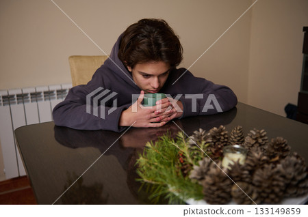Teen Boy Sits At Table With Mug, Reflective Mood, Holiday Pine Decor Nearby 133149859