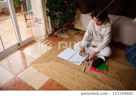 Teenager Studying at Home on a Rug With Notebooks, Pencils, and a Christmas Tree Nearby Teenager Studying at Home on a Rug With Notebooks, Pencils, and a Christmas Tree Nearby 133149880