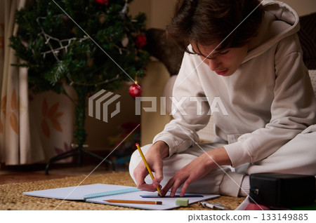 Teenage Boy Drawing On Notebook On Floor Near Christmas Tree In Cozy Home Scene 133149885