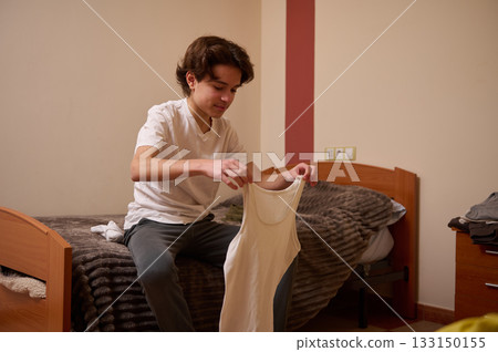 Teenage Boy Folding White Tank Top In Cozy Bedroom, Casual Home Life Teenage Boy Folding White Tank Top In Cozy Bedroom, Casual Home Life 133150155