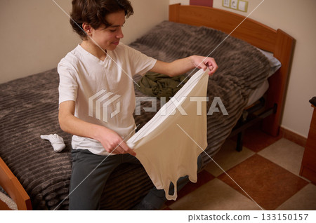 Teen Boy Folding White T Shirt In Bedroom: Casual Daily Life Scene Teen Boy Folding White T Shirt In Bedroom: Casual Daily Life Scene 133150157