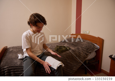 Teenage Boy Sitting On Bed, Smiling While Unfolding Towel In Cozy Bedroom Teenage Boy Sitting On Bed, Smiling While Unfolding Towel In Cozy Bedroom 133150159