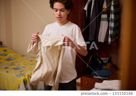 Teenage Boy Folding Clothes in Bedroom With Open Wardrobe, Casual Home Scene Teenage Boy Folding Clothes in Bedroom With Open Wardrobe, Casual Home Scene 133150188