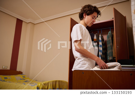 Teen Boy Folding Clothes In Bedroom By Open Wardrobe Teen Boy Folding Clothes In Bedroom By Open Wardrobe 133150193