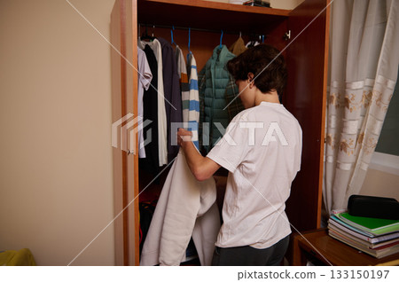 Teen Boy Organizing Closet Clothes and Choosing a Jacket in a Casual Home Scene 133150197