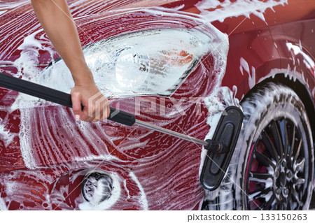 Red car washed in self serve carwash detail on brush moving over front hood above wheels, leaving strokes in white soap Red car washed in self serve carwash detail on brush moving over front hood above wheels, leaving strokes in white soap 133150263