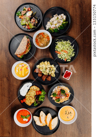 Overhead flat lay of assorted restaurant dishes, soups and salads on wooden table 133150774