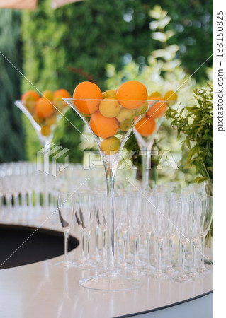 Tall martini centerpiece with citrus above rows of champagne flutes at an outdoor bar 133150825