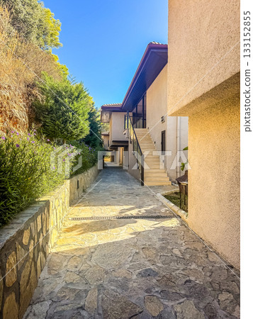 Bright outdoor view of a narrow stone pathway leading to a modern beige house with stairs and greenery around, under a clear blue sky. High quality photo 133152855