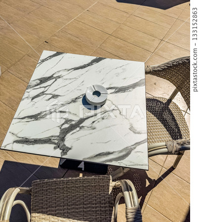 Elegant top view of a marble square table with a white ashtray and wicker chairs on a tiled outdoor patio under bright sunlight. High quality photo Elegant top view of a marble square table with a white ashtray and wicker chairs on a tiled outdoor patio under bright sunlight. High quality photo 133152863