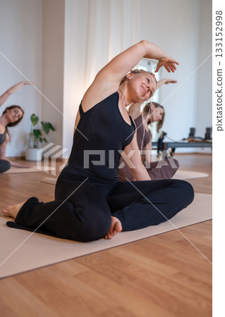 Group of woman carefully doing mermaid exercise, woman engaging core during seated twist in bright studio Group of woman carefully doing mermaid exercise, woman engaging core during seated twist in bright studio 133152998