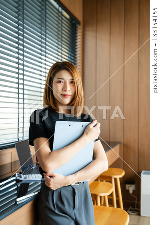 Asian businesswoman in a modern office gives a thumbs-up while holding a clipboard. A confident portrait symbolizing professional success Asian businesswoman in a modern office gives a thumbs-up while holding a clipboard. A confident portrait symbolizing professional success 133154155