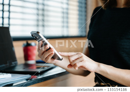 Asian businesswoman using smartphone and laptop in a modern workspace by a window, showcasing digital communication 133154186