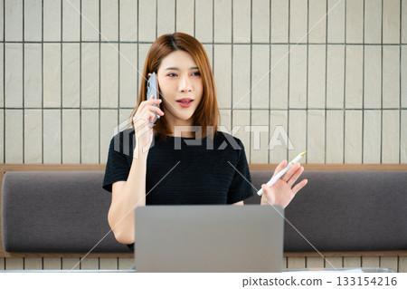 Asian businesswoman talking on smartphone while holding a pen and reviewing a laptop in a modern office 133154216