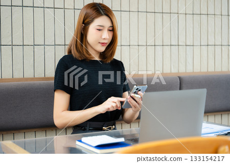Asian businesswoman calculates finances on a clipboard with calculator by her laptop and smartphone in office 133154217