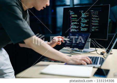 Programmer using stylus and laptop to write code at night. Coding workspace with tablet, monitor, and dark theme 133154306
