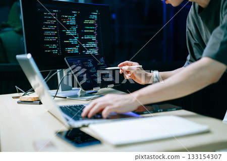 Programmer using stylus and laptop to write code at night. Coding workspace with tablet, monitor, and dark theme 133154307