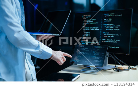 Young male programmer working with tablet and screen displaying code. Modern tech office setup, great for software Young male programmer working with tablet and screen displaying code. Modern tech office setup, great for software 133154334