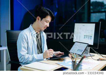 Asian male designer pointing at UX wireframes on screen and tablet in modern office. Ideal for themes in UI UX, digital product design, software 133154340