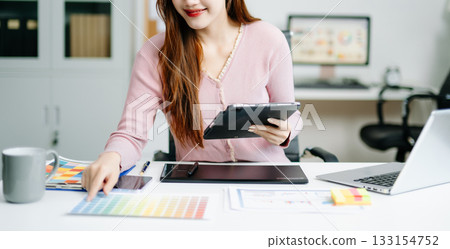 Young graphic designer using pen tablet and laptop at creative desk in modern office. Surrounded by color palettes 133154752