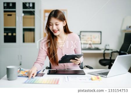 Young graphic designer using pen tablet and laptop at creative desk in modern office. Surrounded by color palettes 133154753