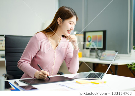 Asian designer using digital pen and laptop in a creative office, showcasing professional work, innovation, 133154763