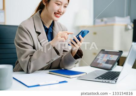 Smiling businesswoman talking on phone while working with laptop in a modern office. Perfect for themes of communication, marketing, finance Smiling businesswoman talking on phone while working with laptop in a modern office. Perfect for themes of communication, marketing, finance 133154816