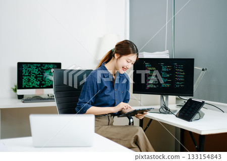 Young female programmer working with tablet and screen displaying code. Modern tech office setup, great for software, coding, IT 133154843
