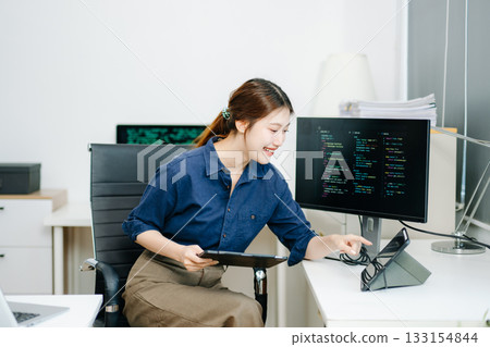 Young female programmer working with tablet and screen displaying code. Modern tech office setup, great for software, coding, IT 133154844