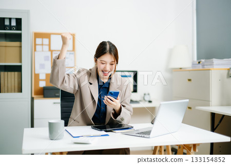 businesswoman celebrating success at her desk with laptop and tablet in a modern office. Perfect for themes of victory, achievement, productivity 133154852