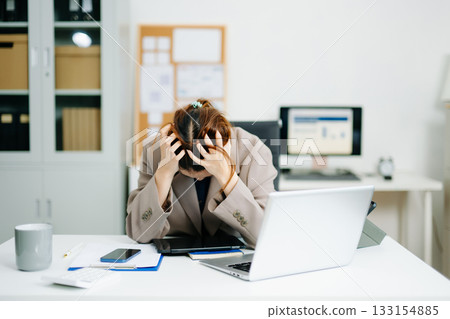Young woman overwhelmed and exhausted at her desk in modern office. Struggling with stress, deadlines, and data reports. 133154885