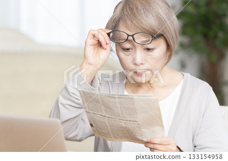Senior woman reading a newspaper wearing reading glasses in the living room Senior woman reading a newspaper wearing reading glasses in the living room 133154958
