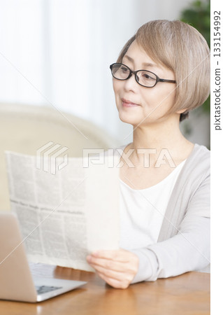 一位老婦人在客廳裡戴著老花眼鏡看報紙。 133154992