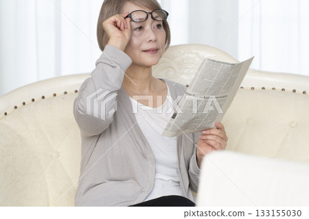 一位老婦人坐在客廳的沙發上，戴著老花眼鏡看報紙。 133155030