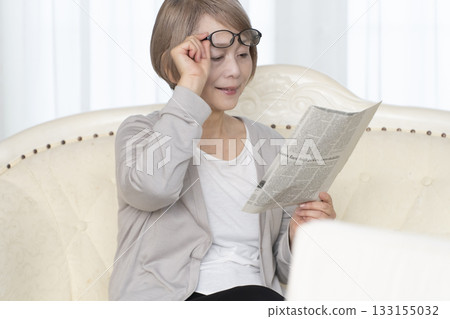 A senior woman sitting on the sofa in the living room and reading a newspaper wearing reading glasses A senior woman sitting on the sofa in the living room and reading a newspaper wearing reading glasses 133155032