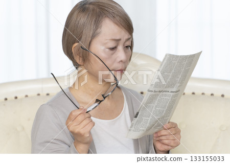 一位老婦人坐在客廳的沙發上，戴著老花眼鏡看報紙。 133155033