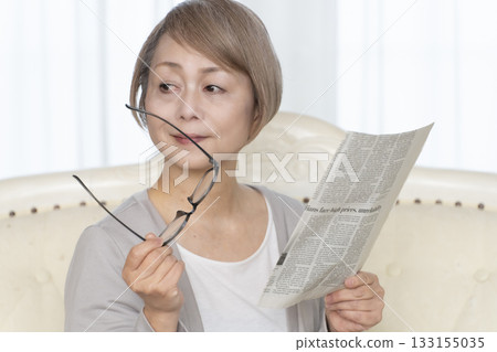 A senior woman sitting on the sofa in the living room and reading a newspaper wearing reading glasses A senior woman sitting on the sofa in the living room and reading a newspaper wearing reading glasses 133155035