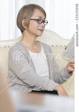 一位老婦人坐在客廳的沙發上，戴著老花眼鏡看報紙。 133155036