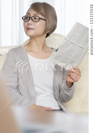 A senior woman sitting on the sofa in the living room and reading a newspaper wearing reading glasses 133155038