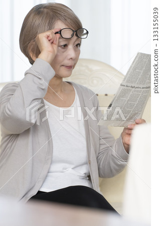 一位老婦人坐在客廳的沙發上，戴著老花眼鏡看報紙。 133155039