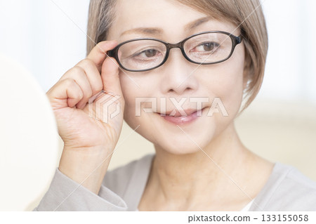 Senior woman wearing reading glasses and looking at a hand mirror in the living room 133155058