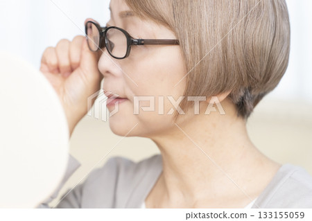 Senior woman wearing reading glasses and looking at a hand mirror in the living room 133155059