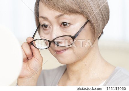 Senior woman wearing reading glasses and looking at a hand mirror in the living room 133155060