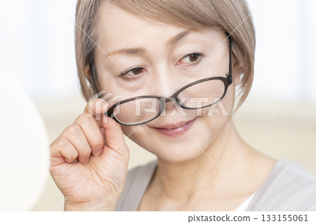 Senior woman wearing reading glasses and looking at a hand mirror in the living room Senior woman wearing reading glasses and looking at a hand mirror in the living room 133155061